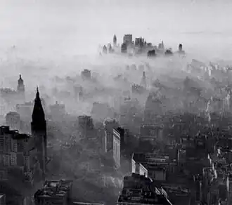 A black-and-white, panoramic view of New York City as seen from a great height. A vast number of buildings and skyscrapers can be seen. A hazy, smoky gas overlays the entire city like a blanket, with a fairly clear skyline only in the far distance at the horizon. Near the closest buildings, the smog appears thin and wispy. The smog appears thicker and thicker around buildings that are farther away from the photographer's position, until shorter buildings near the horizon are almost entirely shrouded and impossible to see under a thick layer of smog. Near the horizon, the clustered tops of tall skyscrapers emerge from within the smog.