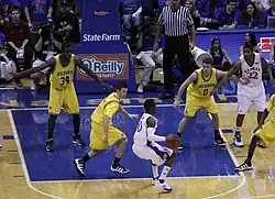basketball players in maize colored uniforms defend against players in white uniforms