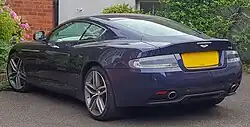 Rear three-quarters view of a very dark purple 2013 facelift DB9 with bushes in the background.