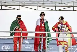 Antonelli wearing his gold medal on the podium of the 2022 FIA Motorsport Games alongside Manuel Espírito Santo and Bruno del Pino, with the Italian flag draped over him