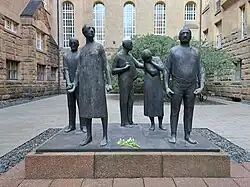 A sculpture of black steel depicting mourning persons. The sculpture is surrounded by an old building.