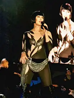 Cher performs on stage in a black bodysuit with sheer panels, jagged embellishments, a short black bob, and a beaded hip scarf. A large screen behind her shows a close-up as she sings.