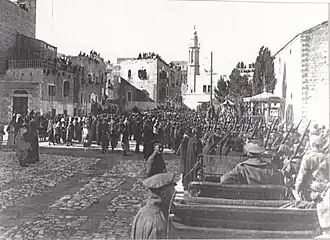 City buildings, some with people looking down from roofs, people and soldiers in a large square, with motor car in foreground