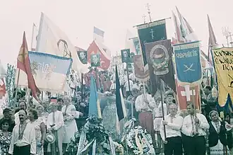 Several people wearing the traditional Ukrainian clothing, vyshyvanka, carry a variety of regional flags and portraits of national leaders as they stand around a headstone adorned with Ukrainian national symbolism.