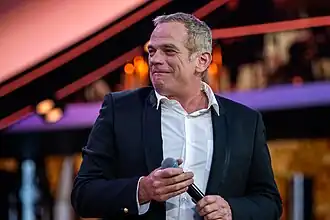 Garou wearing a white shirt and dark jacket, holding a microphone and looking right of camera while grimacing or perhaps near-smiling