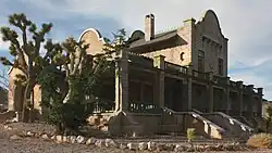 An ornate, two-story masonry building rests under a partly cloudy sky in a setting of gravel, two spiny trees, and many low shrubs. Its windows are boarded.