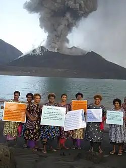 A group of women holding signs supporting gender equality with an erupting volcano in the background across a bay