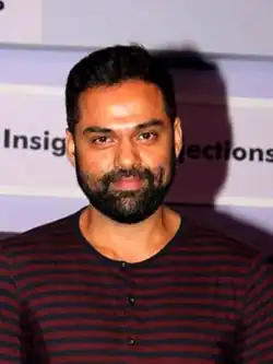 A middle-aged Indian man wearing a black-and-red-striped shirt smiling at the camera. He has short, black hair; a short, face-hugging beard and a moustache.
