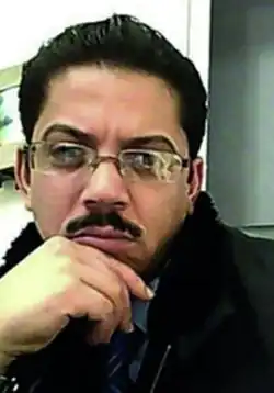 fuzzy internet video image of a middle-eastern male with dark slicked-back hair and moustache in an office setting, and wearing spectacles and a light outdoors jacket
