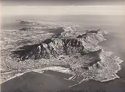 Black-and-white photograph of a city, taken from the air. The city is situated between an ocean and a large flat mountain range.