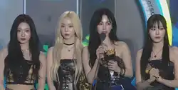 Four women standing on a stage, dressed in matching black and gold outfits and have stylish hairstyles, with one having platinum blonde hair while the others have dark hair. One of them is holding a microphone and a golden award while speaking, while the others stand attentively.