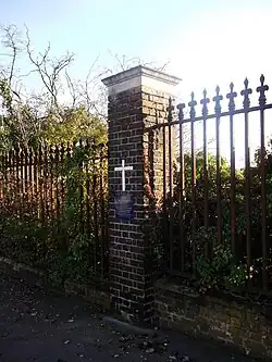 Brick pillar in the middle of iron railings. There is a white cross on the pillar, with a square blue plaque below reading "In Memory of the pilot and seven passengers who lost their lives in the crash of Imperial Airways De Havilland 34 Airliner G-EBBX near the site of this memorial on Christmas Eve 1924"
