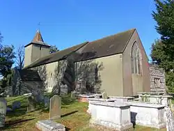 Three-quarter view of a long, low, grey church behind tombs and gravestones in a churchyard. There is a spire-topped tower at the far end of the brown tiled roof.