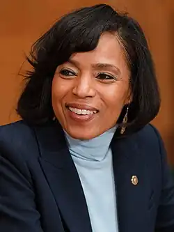 Alsobrooks smiling and looking to her left on a brown background, wearing a light blue shirt and black jacket with a U.S. Senate lapel pin.