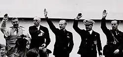Black-and-white photograph of the upper bodies of five white men. Four raise their right arms in a fascist salute. From left to right: a man in bishop robes; a man in a civilian suit with his right hand reaching for his breast pocket; a man in a dark uniform; a man with glasses in a military uniform and a man in a dark uniform (Suanzes).