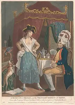 In an elaborately decorated eighteenth-centiry bedroom, a woman stands in an assertive pose wearing a man's tricorne hat, facing a man who sits submissively wearing a large feminine wig and holding a fan.
