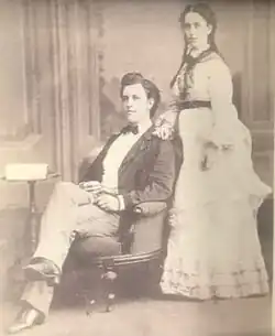 Black and white of a middle-aged man and woman. The man has dark hair, is seated in an arm chair, wearing a dark-colored suit jacket, light-colored pants, white dress shirt and black bow tie. The woman is standing behind and to the side of the chair, wearing a full-length white dress with a dark-colored belt, and dark hair braided into two ponytails.