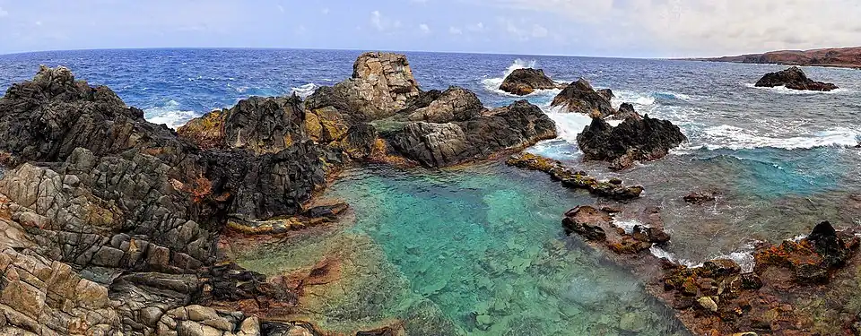 Photo of a rocky shoreline, with a natural pool filling up with clear, blue-green water from the sea. Waves crash close by, and the blue sea lies beyond.