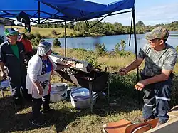 A man turns a spit with chochoca on it, while a woman brushes it, presumably with lard