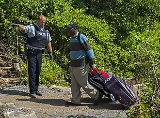 A bald uniformed police officer wearing black gloves and a blue flack jacket with "POLICE" written on it in white letters at left points in that direction while a man on the right in a blue and brown striped sweater wearing a baseball cap pulls a wheeled suitcase behind him on a dirt pathway with shrubs behind him