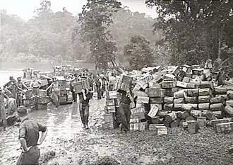 Black and white photo of a large number of small wooden crates stacked on muddy ground, with men wearing military uniforms carrying additional small wooden crates