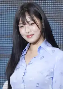 Woman with long, dark hair and bangs, smiling gently at the camera. She is wearing a light blue button-up shirt with a subtle pattern.