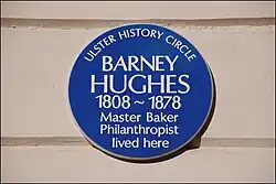 "Circular blue plaque with white lettering erected on a cream wall. Wording is as follows: 'Ulster History Circle. Barney Hughes 1808 ~ 1878, master baker, philanthropist, lived here'"