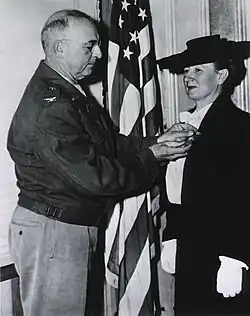 3/4 length standing portrait of a woman in a suit, gloves, wearing a large hat, being pinned with a medal by a man dressed in a military jacket.