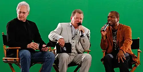 Shatner, Spiner and Burton are speaking together at Comic-Con. They are all sitting in direction chairs in front of a green screen.