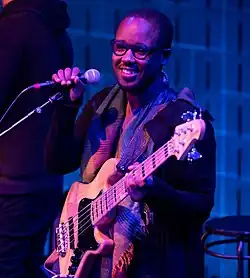 Louis Cato standing onstage with an electric bass guitar, holding a microphone in a stand and grinning at audience