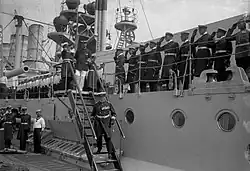 Men in military uniforms walk down stairs on a large warship as dozens of sailors look on and salute.