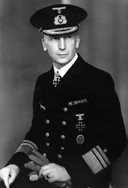 A man wearing a peaked cap and dark military uniform with an Iron Cross displayed at the front of his uniform collar. He is holding gloves in his hands.