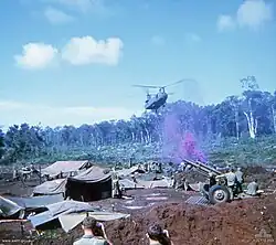Colour image of a large two-rotored helicopter flying past a line of trees and preparing to land in an open area cleared of vegetation. In the foreground are a large number of tents, hootchies and ground sheets, while some soldiers are standing around an artillery piece. Purple smoke is rising from the position.