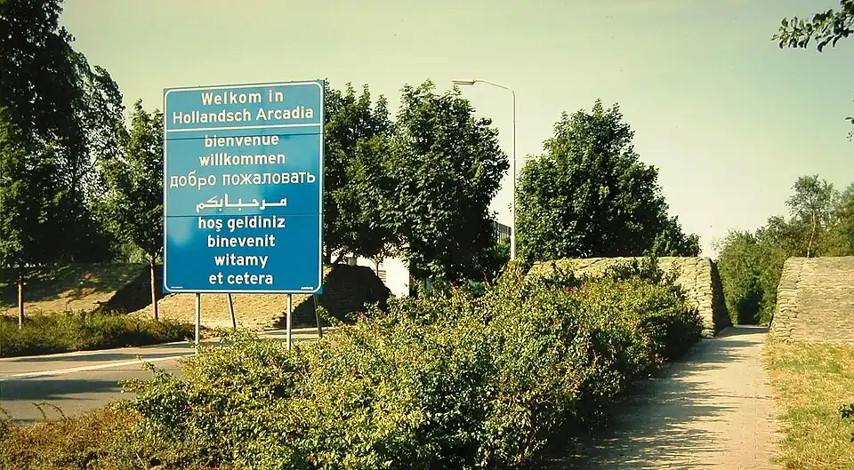 "Welcome" highway sign in several languages, next to a footpath through a wall of sandbags