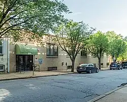 Photograph of a street with a slight slope. Across the road, there is a one-story building in yellow brick. The glass door is covered by a green canopy with the number 1209. There is a logo with "CT" on the door. Along the pavement there is a park meter, four trees and three dark parked cars.