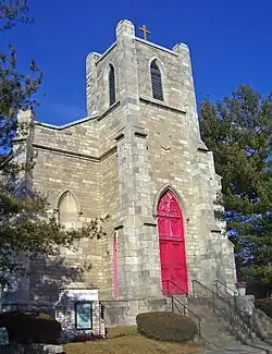 A narrow light brown stone building with a red door in a pointed arch. Above it is a square tower with a cross on top. A small sign in front says "Calvary Baptist Church".
