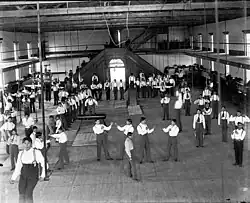 Carlisle Students in School Uniform Exercising Inside Gymnasium; Some with Indian Clubs, Others with Gymnastic Equipment; Non-Native Group Watching, 1879