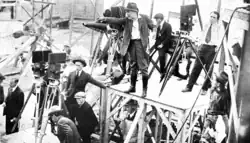 DeMille on a stand, holding a megaphone, giving commands to crew members which surround him and several film cameras