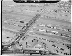 An aerial view of a rail yard, in which a number of freight train cars are visible, predominantly boxcars. A road bridge crosses over the rail yard and connects to a highway in the background.