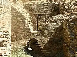 Inside daytime view of a ruined and ceiling-less rectangular room. Tawny-beige stacked sandstone bricks compose walls rising from brush-covered ground. The several walls visible in the image are up to perhaps a dozen feet in height. In the wall immediately at center, a triangle-shaped entrance several feet high leads to an adjacent chamber behind. The upper part of the same wall, shaped like an inverted-triangle, has fallen away or otherwise been removed, revealing a rectangular doorway leading to yet another concealed room. At left and right are two similar walls perpendicular to the one at middle.