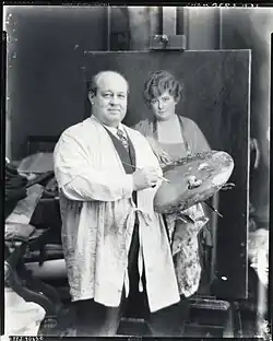 Black and white photograph of a man in a painter's smock holding a palette, standing in front of a painting