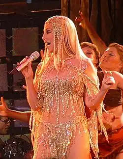 Cher sings on stage in a shimmering gold beaded outfit with fringe, a matching headpiece and long platinum blonde hair, surrounded by backup dancers.
