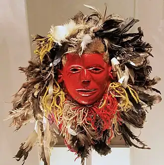 A red mask adorned with feathers, fur, and rope around the edges, not dissimilar to a lion's mane.