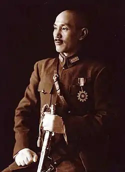 A Chinese man in military uniform, smiling and looking towards the left. He holds a sword in his left hand and has a medal in the shape of a sun on his chest.
