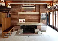The fireplace at the eastern end of the living room. The mantelpiece is made of stone, and the fireplace is flanked by brick columns. There is a gap between the columns above the fireplace, and the dining room is visible through the gap.