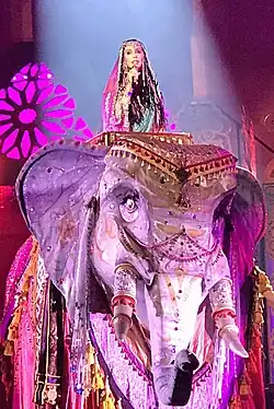 Cher wearing an elaborate bejeweled costume with a veil, singing into a microphone while seated atop a large, ornately decorated mechanical elephant during a stage performance.