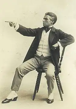 A young Claude Rains dressed as a lawyer, sitting on a chair and pointing
