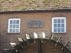 Brick building with tiled roof and two square windows, below which is a water wheel. Between the windows there is a wooden plaque, which reads "Cobham Mill Restored 1993 by Cobham Mill Preservation Trust and the National Rivers Authority".