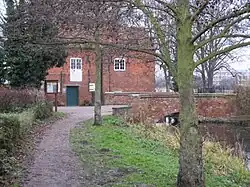A view of the mill building from the footpath on the north bank. The pool and the bridge across the race are visible to the right, but the red brick face of the building dominates. In the extreme foreground on the left is a bare-branched tree, its trunk lightly flecked with lichen. In the middle distance on the left is a holly, dark green and dense.