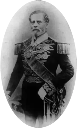 Tintype photograph depicting a full-length portrait of an older, bearded man wearing an elaborately embroidered military dress uniform with heavy epaulets, sash of office, sword, gloves and holding a bicorn hat in his right hand
